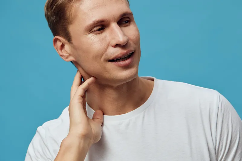 Close-up portrait of a smiling young Caucasian man with short brown hair wearing a white t-shirt against a plain blue background, expressing friendly and approachable emotions. Studio shot with natura