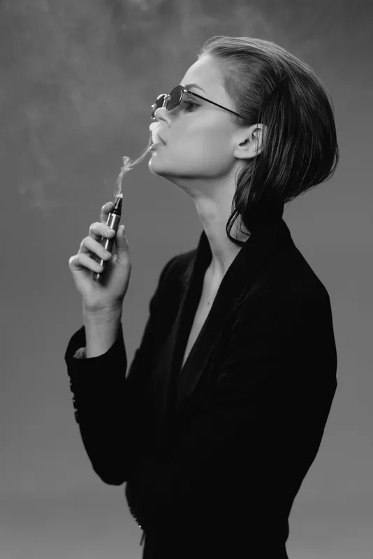 Black and white artistic portrait of a stylish woman smoking with sunglasses and slicked-back hair, expressing confidence and attitude.