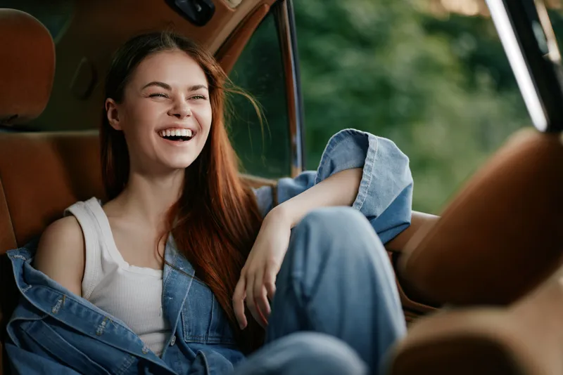 Young woman laughing in a car, wearing denim clothing and a joyful expression, outdoors, surrounded by greenery, showcasing a carefree lifestyle and seasonal warmth.