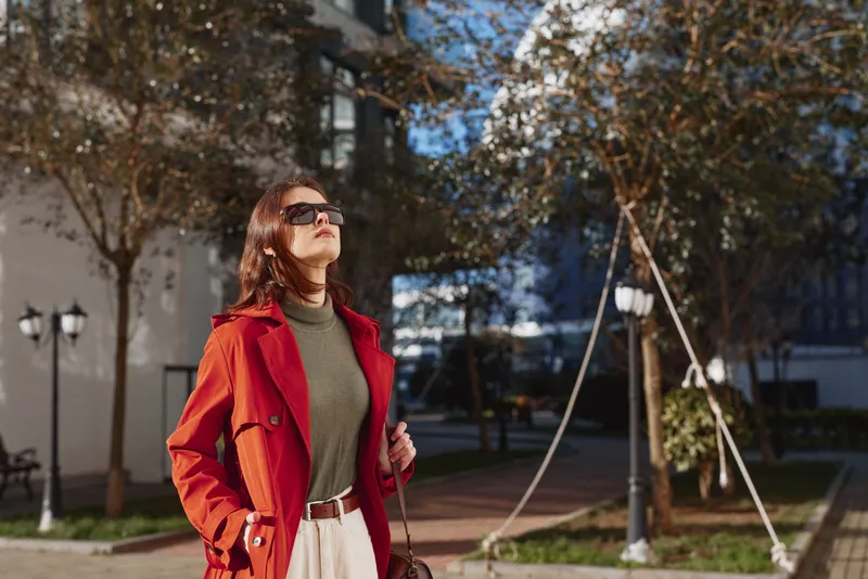A confident woman in a red coat and sunglasses enjoys a sunny day outdoors, standing on a city street with autumn trees and buildings in the background.