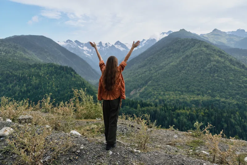 A traveler stands on rocky ground with arms raised overlooking a vast mountain landscape and valley evoking freedom adventure and a deep connection with nature