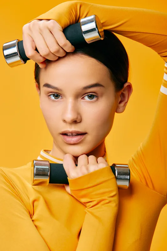 Female fitness model lifting light dumbbells, stretching arms in a bright orange workout top, posed against a vibrant yellow studio backdrop.