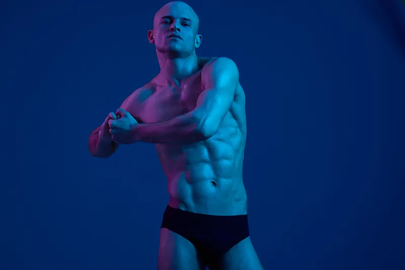 Athletic man posing in dramatic lighting, showcasing a strong physique and confident demeanor. The blue and purple tones enhance the visual impact of the image.
