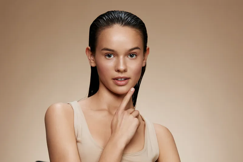 Young female model with wet hair poses thoughtfully against a neutral beige background, showcasing natural beauty and confidence in a minimalistic style