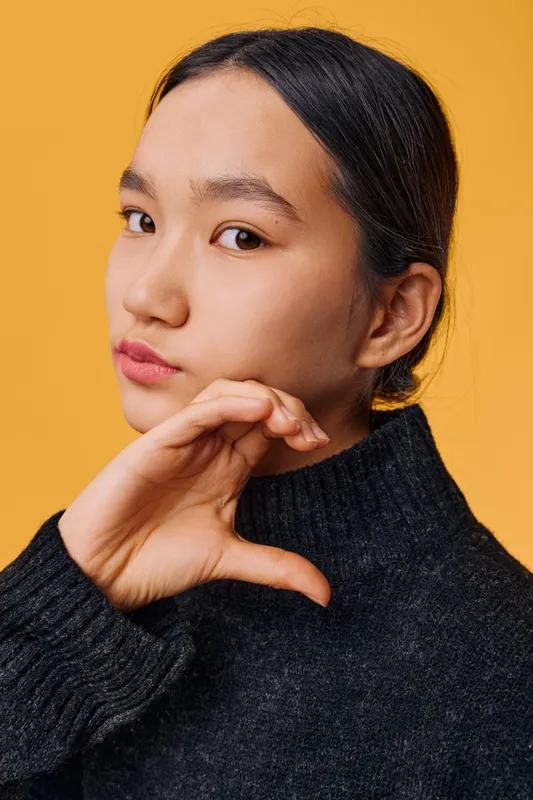 Portrait of young woman with clear skin and dark hair making heart shape with hand on yellow background, wearing black sweater, showing emotion and gesture of love and care.