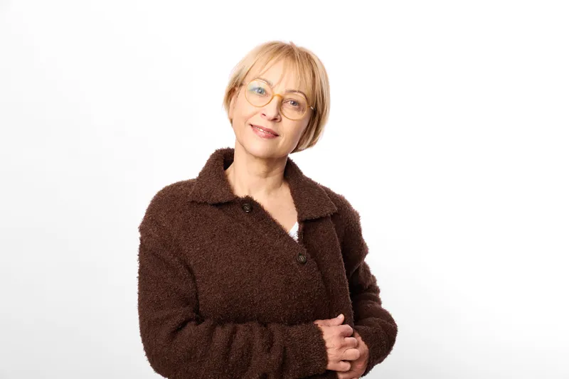 Mature woman with glasses and brown coat smiles with arms crossed in studio portrait, isolated on white background, calm confident expression, copy space