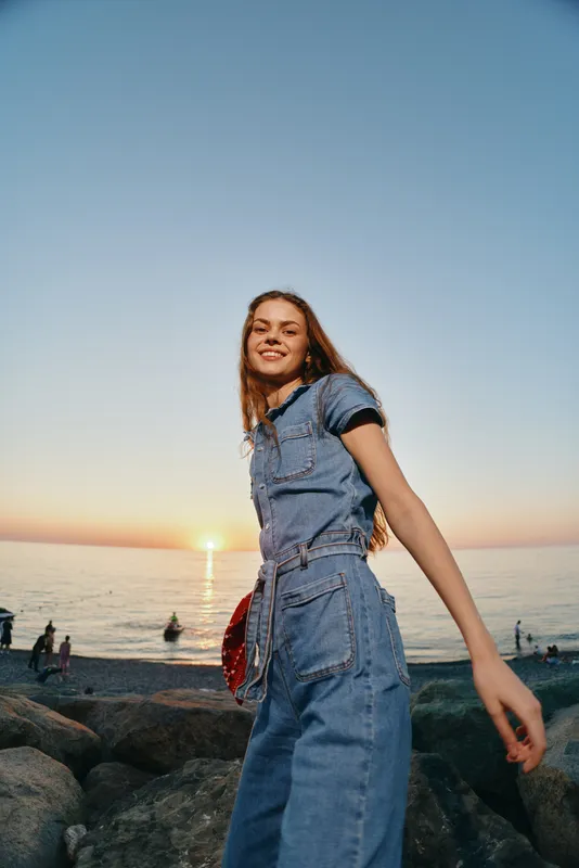 Lifestyle woman in modern boho-western denim street style smiling outdoors by the beach at sunset, with warm film color and relaxed confident mood