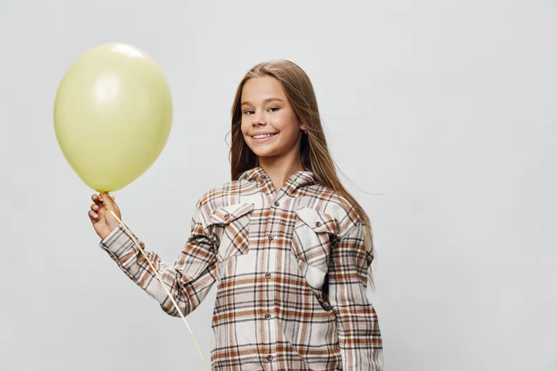 Girl smiling and holding yellow balloon in hand. Child in casual plaid shirt posing with party decoration on light background. Happy kid with long hair and festive mood.