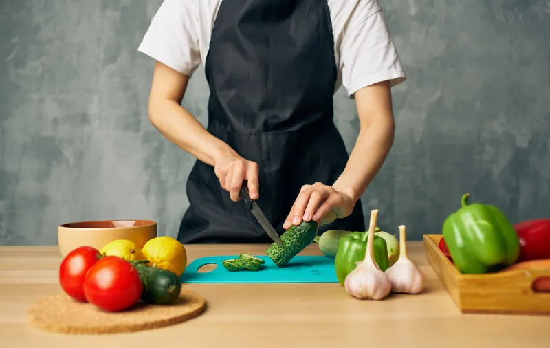 Женщина-повар в черном фартуке нарезает продукты на разделочной доске на кухне