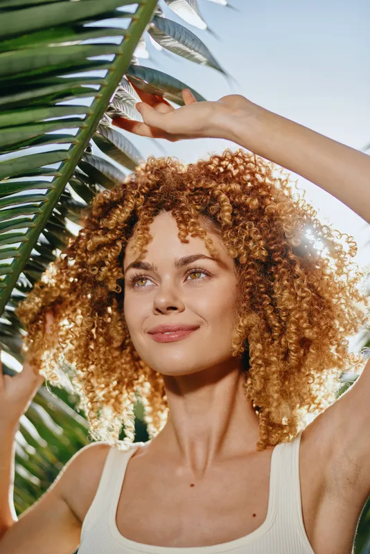 smiling woman with curly hair holds tropical palm leaf under bright sunlight, fresh summer vibe, natural beauty in casual white tank top outdoors