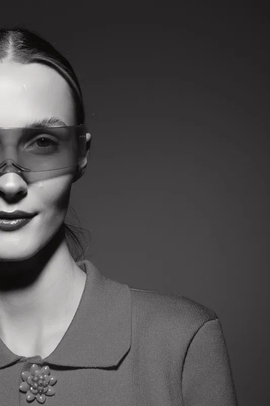 Portrait of a young white woman with fair skin wearing transparent glasses and a textured shirt with a decorative brooch, studio black and white lighting.