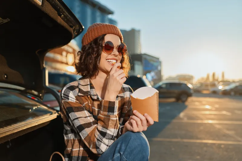 Woman sits at car trunk in parking eating fries, beanie and sunglasses on, smile warm in sunlight. Candid lifestyle shot with authenticity, golden hour glow and emotional storytelling.