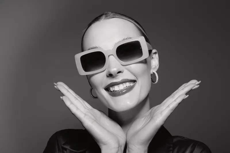 Happy young white woman with pale skin and sunglasses smiles broadly, posing with hands near face against plain grey background. Concept of joy, fashion, and style.