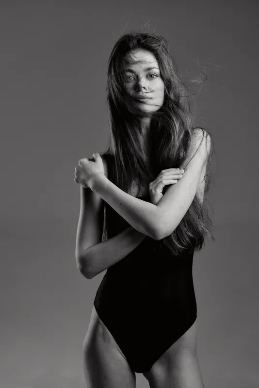 Black and white portrait of a young woman with long hair wearing a black bodysuit, expressing calm and mystery with crossed arms.