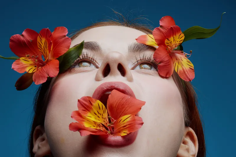 Woman with flowers in her mouth, vibrant colors, blue background, emotional expression, nature inspired beauty concept, highlighting individuality and creativity