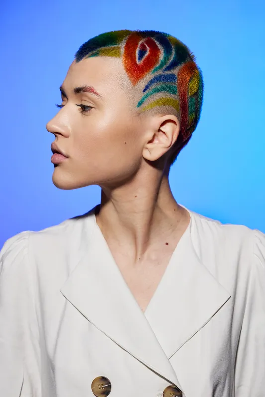 Young woman with colorful shaved hair design in abstract patterns, wearing white blazer, against vibrant blue background, showcasing bold artistic hairstyle.