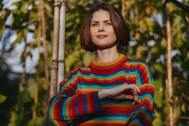 Woman in a colorful striped sweater with short brown hair smiling in a sunlit garden, casual outfit leaning on bamboo stake, relaxed confident expression for lifestyle fashion portrait.