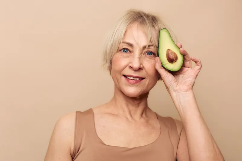 Smiling mature woman with short blonde hair holds half of fresh avocado near face. Beige background and casual outfit create natural wellness and healthy lifestyle concept for skincare and diet.