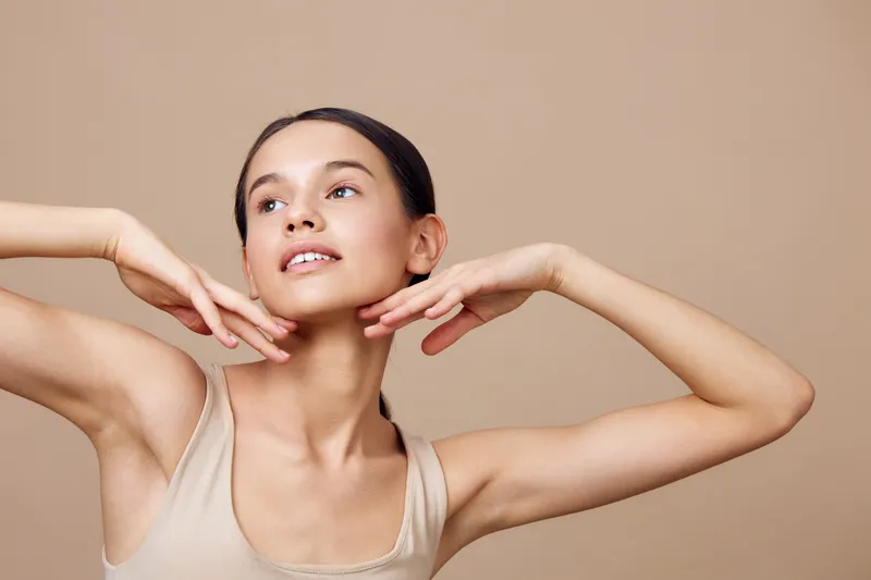 Young woman posing gracefully against a neutral background, exuding confidence and joy, wearing a light beige outfit that complements her warm skin tone