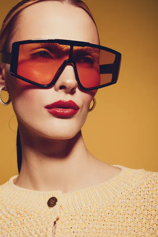 Stylish woman with oversized orange sunglasses and red lipstick poses against yellow background. Fashion portrait with modern accessories and confident expression in studio lighting.