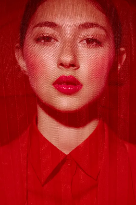 Portrait of a young woman with bright red lipstick and intense gaze behind red sheer fabric. Creative beauty fashion concept showcasing vibrant colors and bold styling effects.