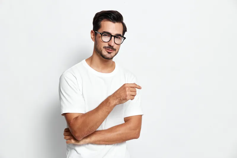 Handsome young man wearing glasses and casual white t-shirt poses confidently while pointing with his finger, isolated on plain white background. Lifestyle concept.