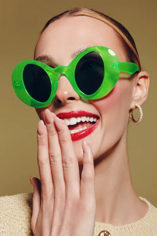 Smiling woman with bright red lipstick wears large green sunglasses and gold hoop earrings. She covers her mouth with hand, posing against beige background. Concept of fashion and style.