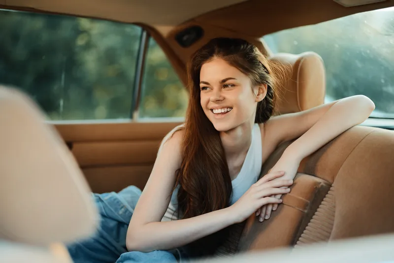 Young woman with long hair smiling in a car, wearing casual attire, capturing a moment of joy and freedom in a serene outdoor setting.