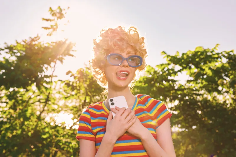 A woman in a striped shirt and retro sunglasses stands outdoors, smiling while holding a smartphone. Bright sunlit scene with vibrant color palette conveys carefree summer lifestyle.