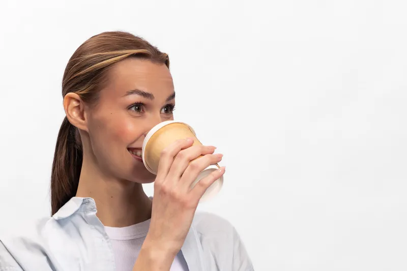 Smiling young woman with brown hair drinks coffee from a paper cup on white background. Concept of morning routine, relaxation, and beverage enjoyment.