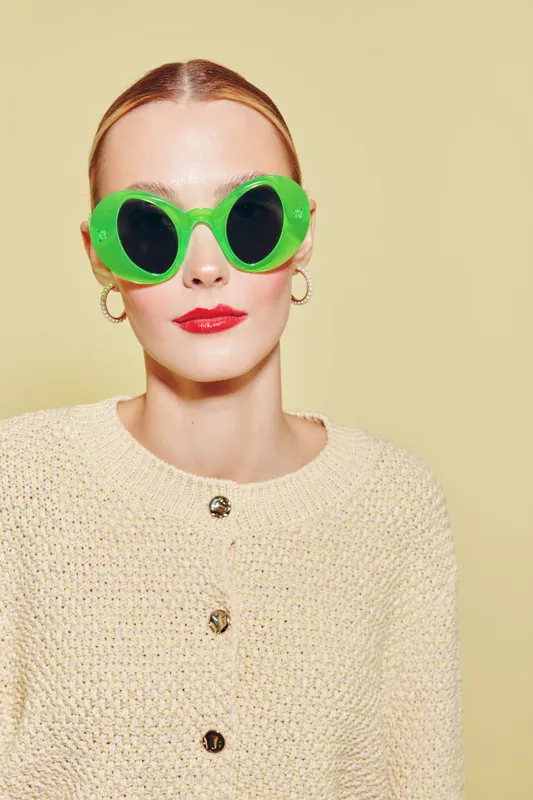 Young woman with slicked back hair wears oversized green sunglasses and red lipstick. She poses against a beige background wearing a textured cream sweater with buttons. Fashion portrait concept.