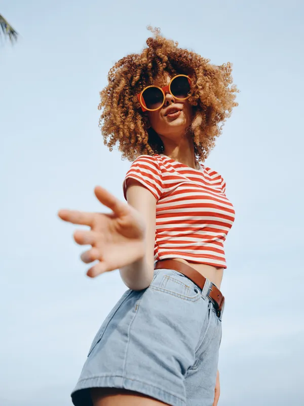 Vibrant portrait of a stylish model with curly hair, wearing a striped red and white crop top and denim shorts, captured outdoors in bright sunlight with bold sunglasses and confident pose.