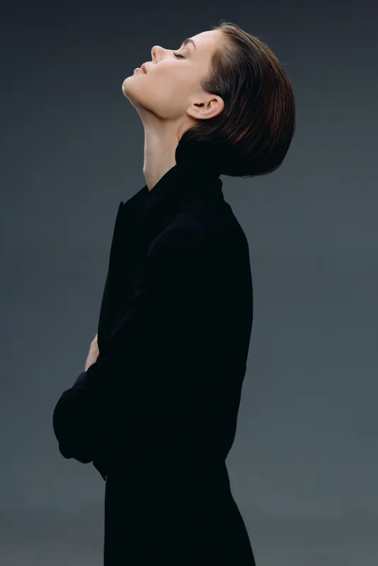 Profile of a thoughtful woman in black clothing with eyes closed against a dark gray background.