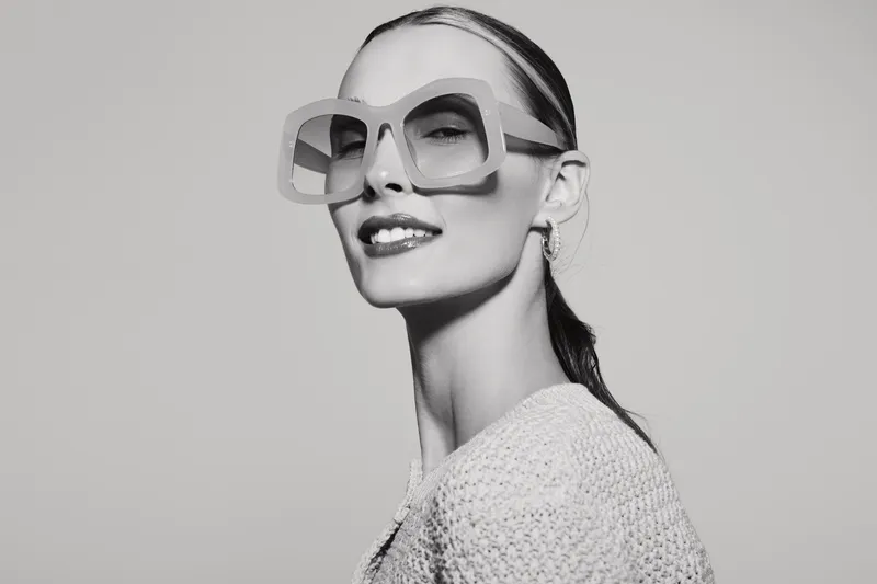 Tall slim caucasian woman with pale skin wears large glasses and knitted top looks confident in studio portrait.