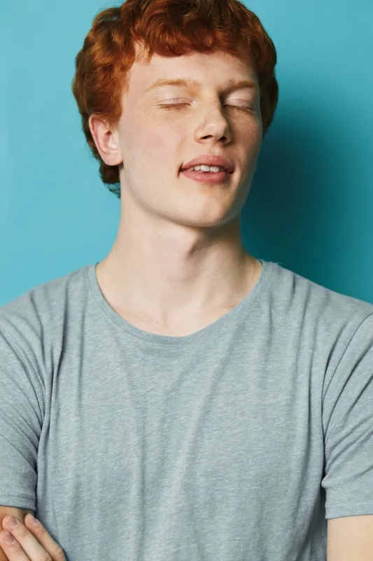 Young man with red hair wearing a gray t shirt standing with crossed arms and closed eyes on blue background. Portrait of relaxed male model with fresh skin and casual style.