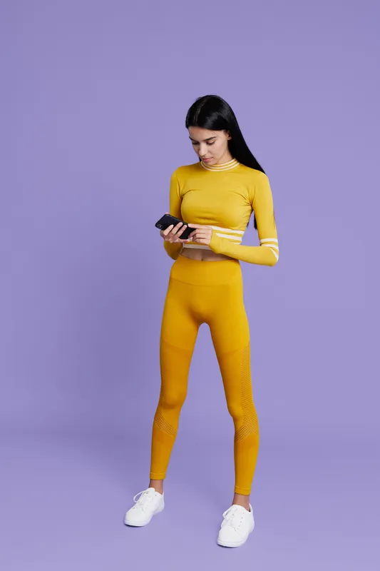 Woman sport fitness wearing bright yellow activewear and white sneakers, standing against purple background, checking smartphone, focused and healthy lifestyle.