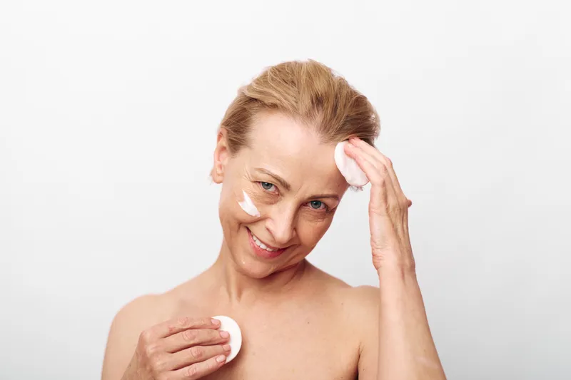 Mature woman with short hair applies facial cream with cotton pads on clean skin. Skincare routine concept with natural beauty and self-care isolated on white background.