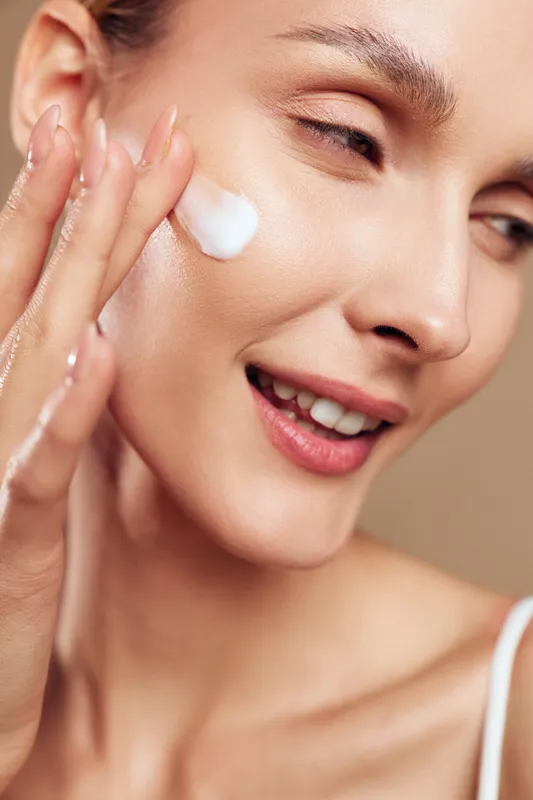 Young woman with light skin applies a dollop of white face cream to her cheek, smiling softly as soft studio light highlights smooth skin against a warm beige background.