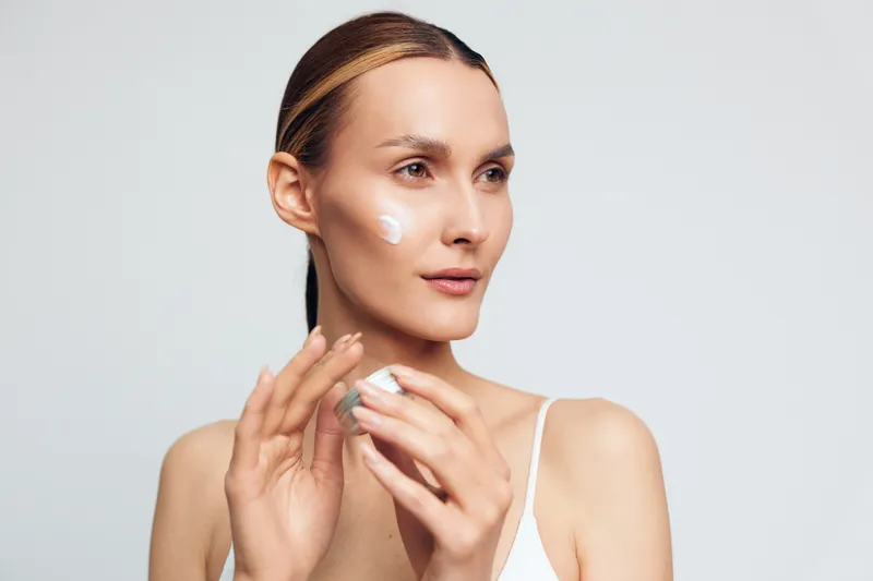 Young woman applying facial cream on clean skin, skincare routine, natural beauty, soft lighting, minimal makeup, neutral background, focused expression, healthy lifestyle