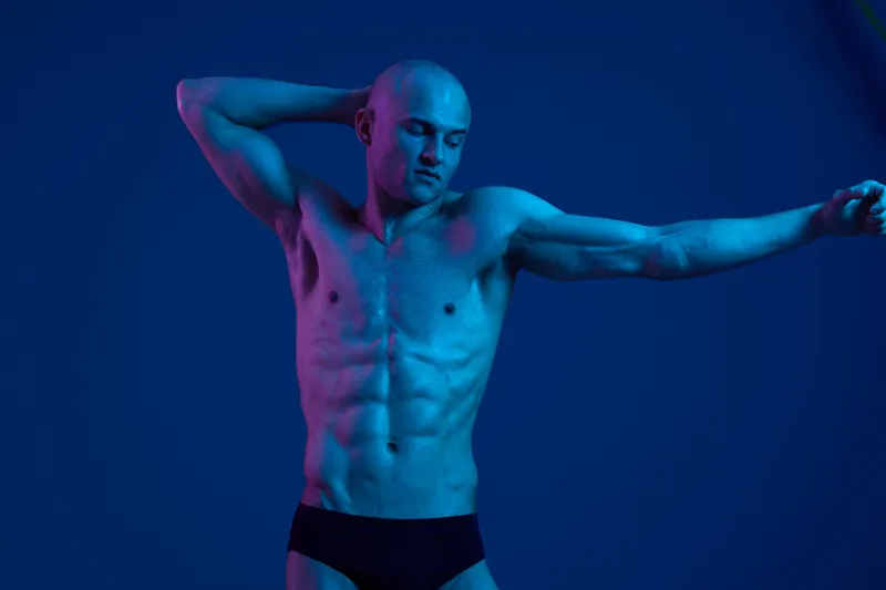 Athletic man posing in vibrant lighting, showcasing strong physique and confident expression. The image captures a playful and dynamic moment in a visually striking setting.