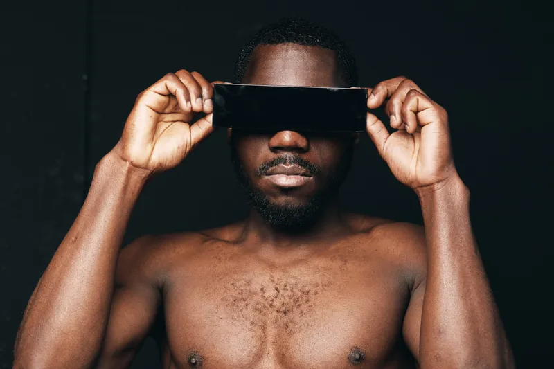 African man with dark skin and bare chest holding black smartphone over eyes in creative conceptual visual style on black background for technology and identity.