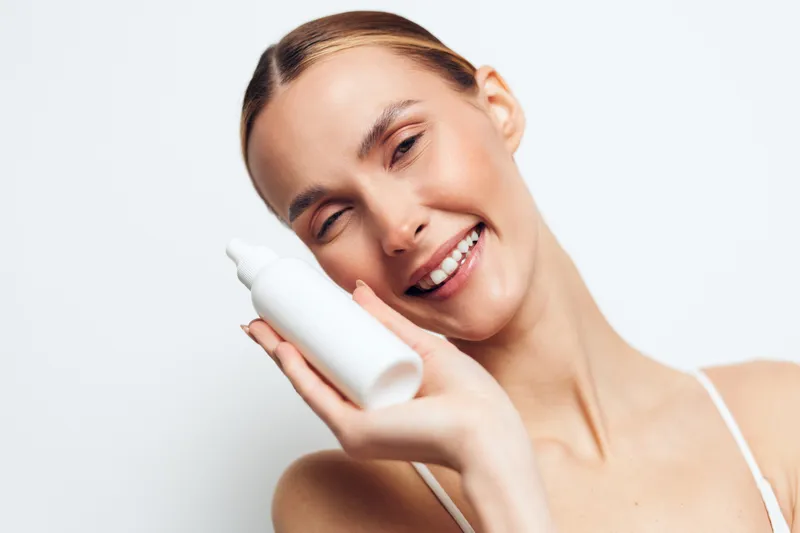 Smiling woman holding white cosmetic bottle near face, clear skin, natural makeup, bright studio background, skincare concept, beauty product presentation.
