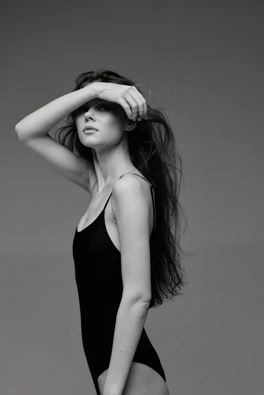 Black and white portrait of a woman with long hair covering her eyes, wearing a black tank top against a plain background.