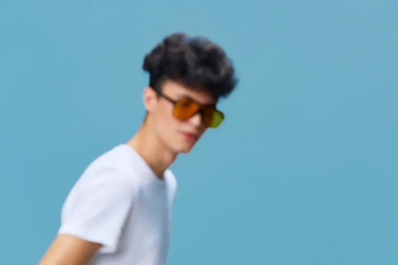Young man wearing sunglasses and a white t-shirt against a blue background, styled with curly hair and casual summer look.