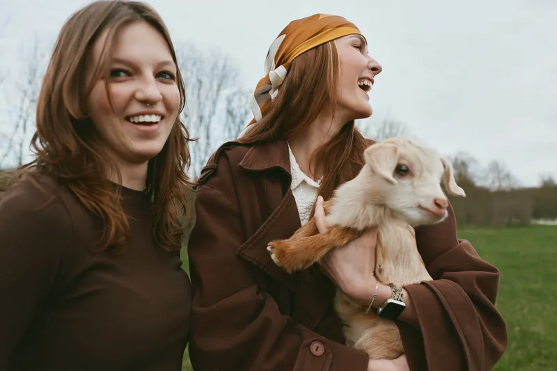Two young women enjoy a joyful moment outdoors as one holds a baby goat while they smile and laugh in casual autumn clothing under an overcast sky.