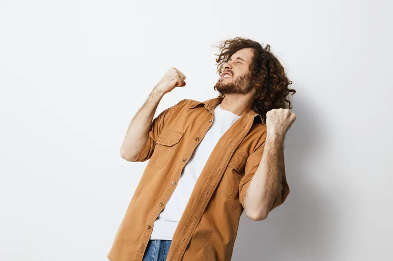 Happy young man celebrating success with joyful expression, wearing casual shirt and t shirt against a neutral background, showcasing positive emotions and energy