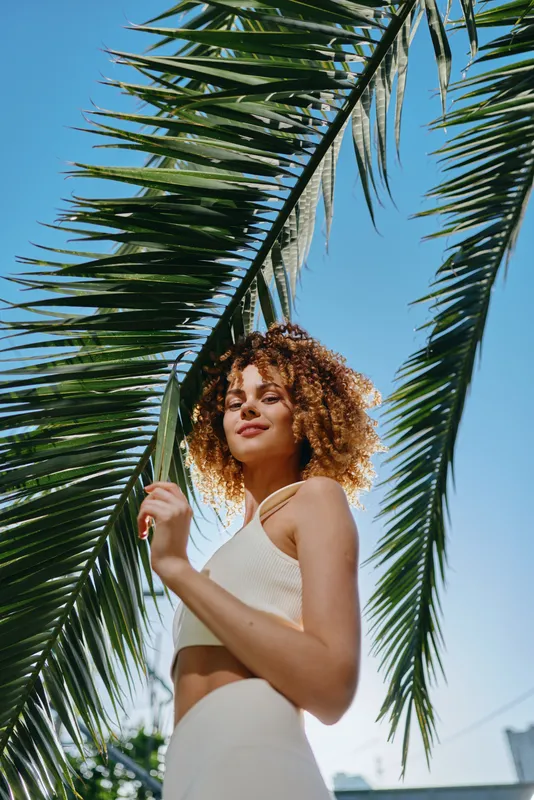 smiling woman with curly hair stands under large palm leaves in bright sunlight, wearing white sleeveless top and light background, tropical outdoor scene