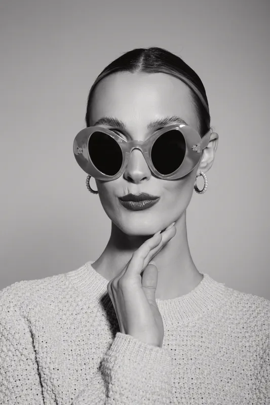 Elegant white woman with fair skin wears oversized round sunglasses and knitted sweater, posing in studio.