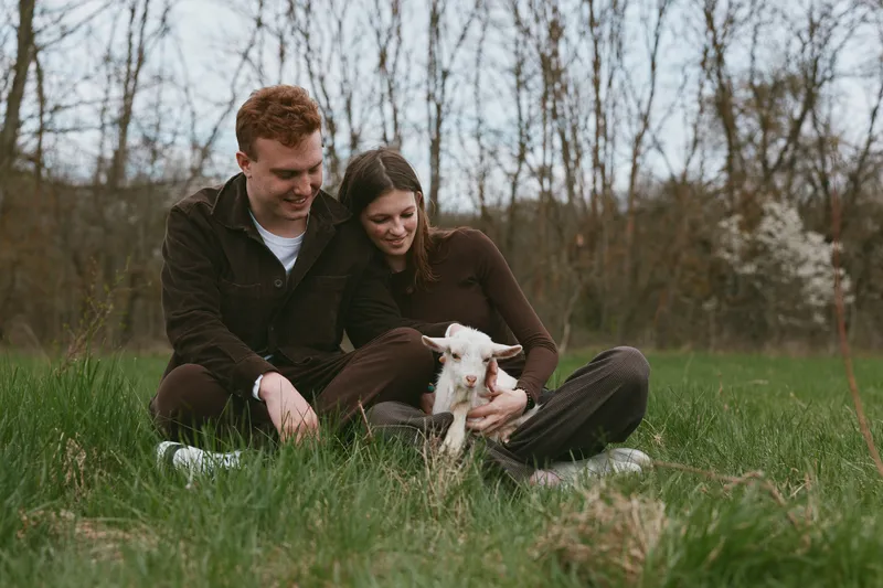 Couple sits on the grass in a park, smiling as they pet a friendly goat kid, enjoying a peaceful outdoor moment together and a touch of rural charm.