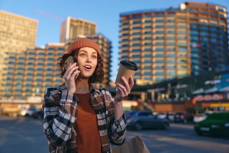 Woman coffee phone city beanie smiling urban scene with plaid jacket, candidly chatting while holding takeaway cup. Authenticity, candid lifestyle, golden hour glow, mindful living, emotional